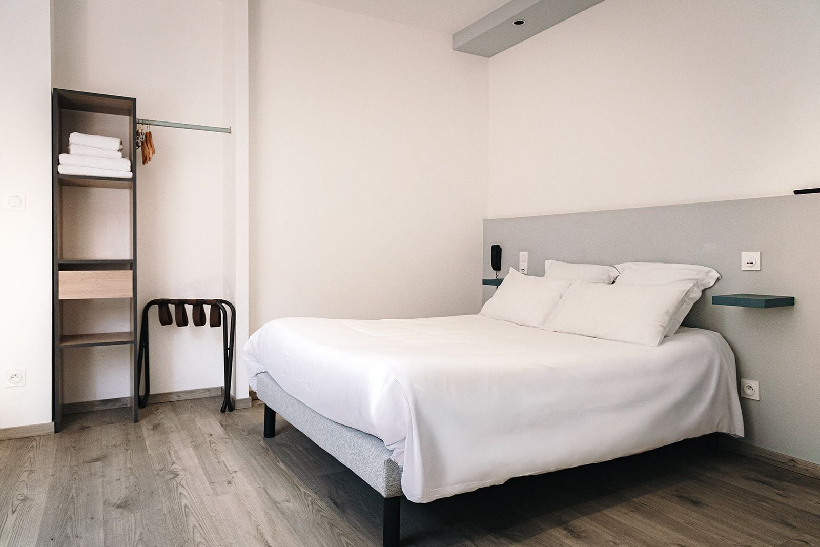 A neatly arranged hotel room with a bed, a shelf with towels, and a chair.