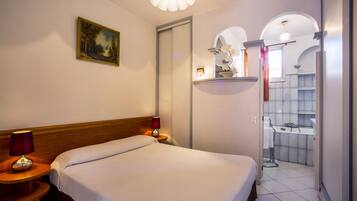 Een hotelkamer met een bed, een nachtkastje met lamp, een schilderij aan de muur, en uitzicht op de badkamer met wastafel en spiegel.