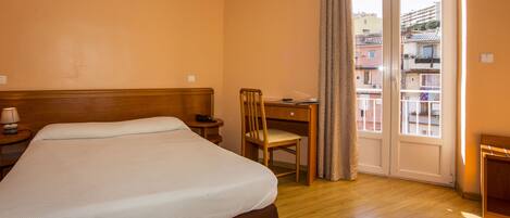 A hotel room with a bed, a desk, a chair, a lamp, and a view of the outside balcony.
