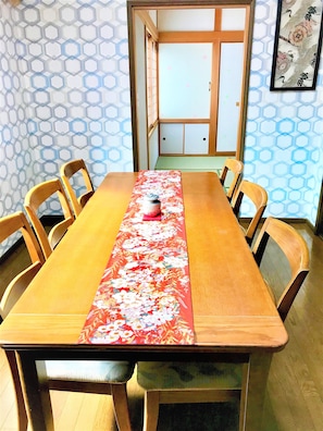 A wooden dining table with a floral table runner, surrounded by wooden chairs. A wooden door and a framed artwork are visible in the background.