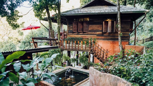 A wooden house with a wooden staircase leading to a deck, surrounded by lush greenery and a small pond. - Taloh Homestay Chiangmai (Mae On)