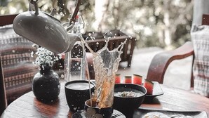 A hand pouring water from a kettle onto a cup, creating a splash. A bowl of food and a plate with eggs are on the table. - Taloh Homestay Chiangmai (Mae On)