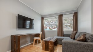Een moderne woonkamer met een flatscreen televisie, een houten bankje, een grijze bank en uitzicht op een woonwijk door grote ramen.