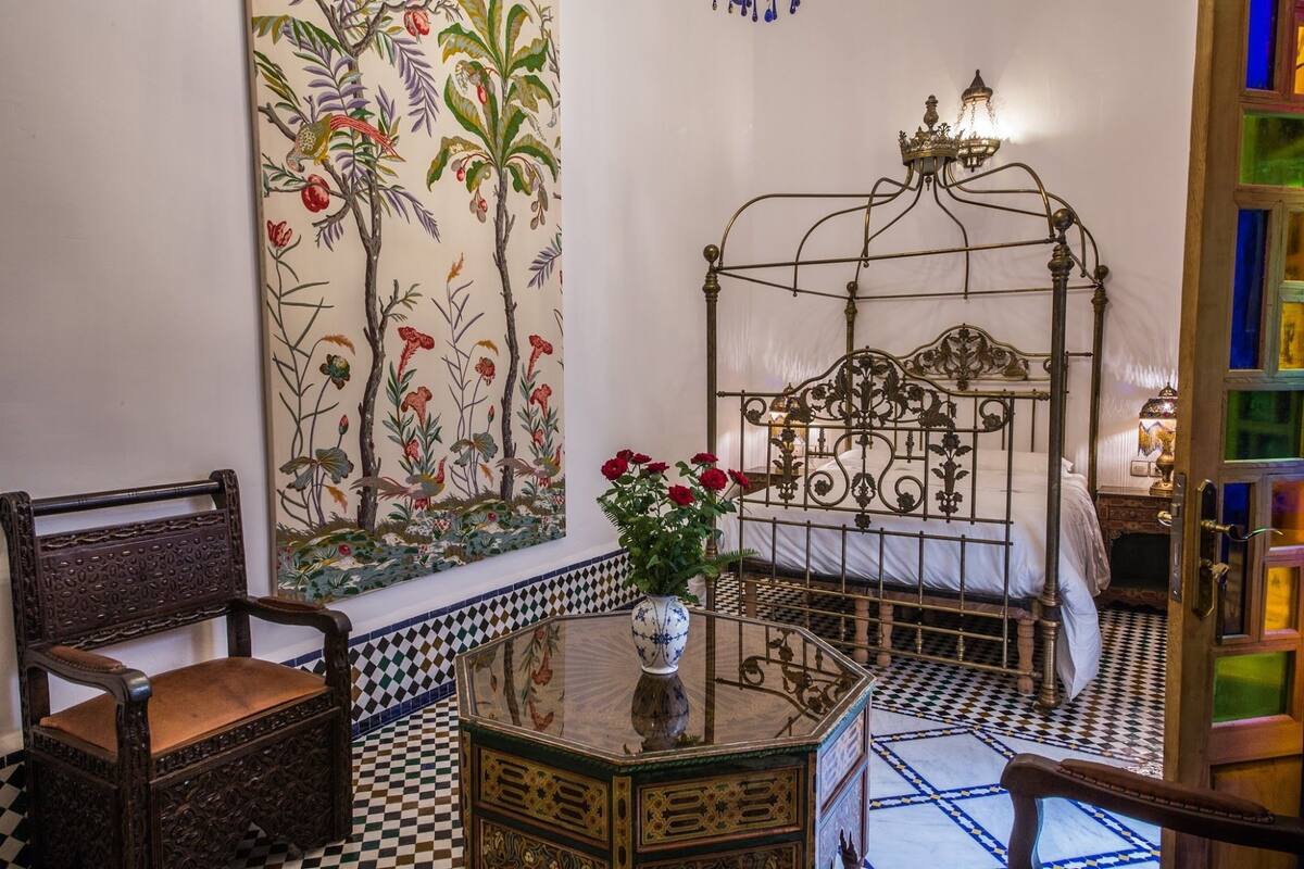 A room with a checkered floor, a metal bed frame, a large floral painting, a wooden chair, and a vase with flowers.