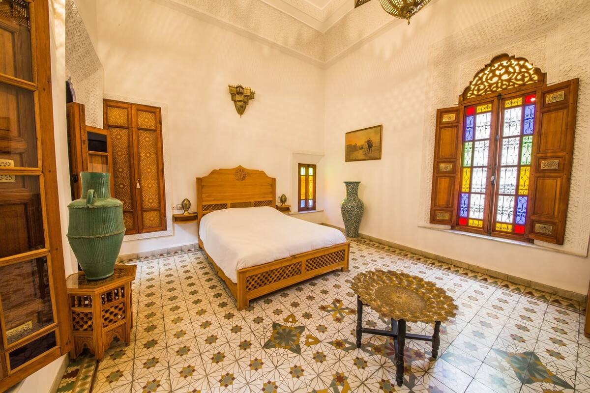 A room with a bed, a decorative chair, a large vase, and a stained glass window.