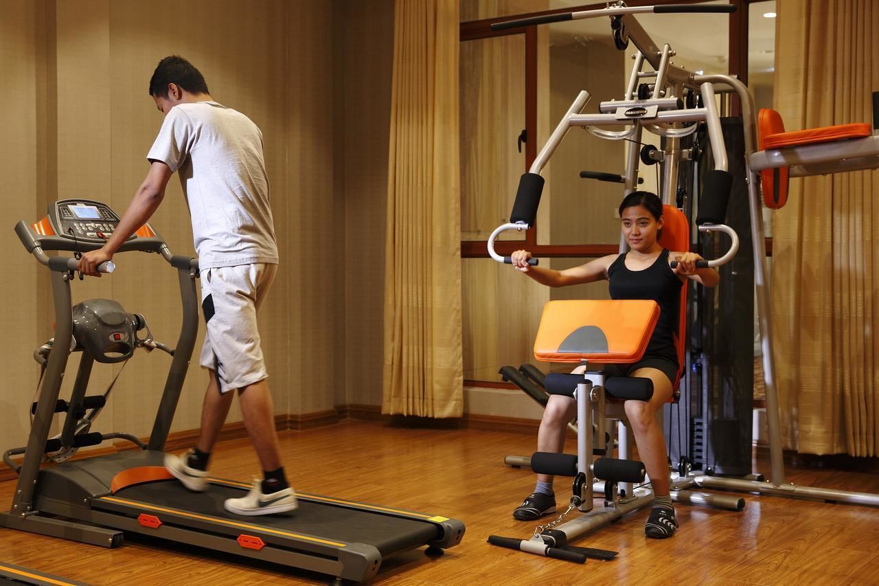 A man on a treadmill and a woman using a leg press machine in a gym with wooden floors and a mirror.