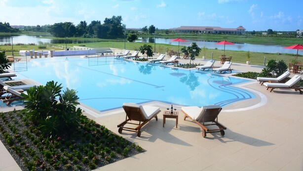 Piscina com espreguiçadeiras e guarda-sóis, cercada por vegetação e com vista para um edifício do outro lado da água.
