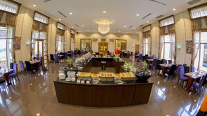 Um amplo hall de hotel com área de buffet central, diversos espaços de estar e grandes janelas que permitem entrada de luz natural.