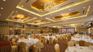 Uma sala de banquetes com mesas redondas, cadeiras douradas, lustres e um palco com um banner.