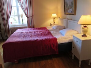 A neatly made bed with a red blanket, two bedside lamps, a white nightstand, and a window with curtains. - Tössestugan Herrgårdshotell (Tösse)