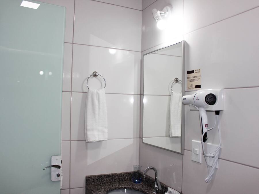Un baño con mampara de vidrio, lavabo con encimera de granito, secador de pelo montado en la pared y toalla colgada en un perchero.