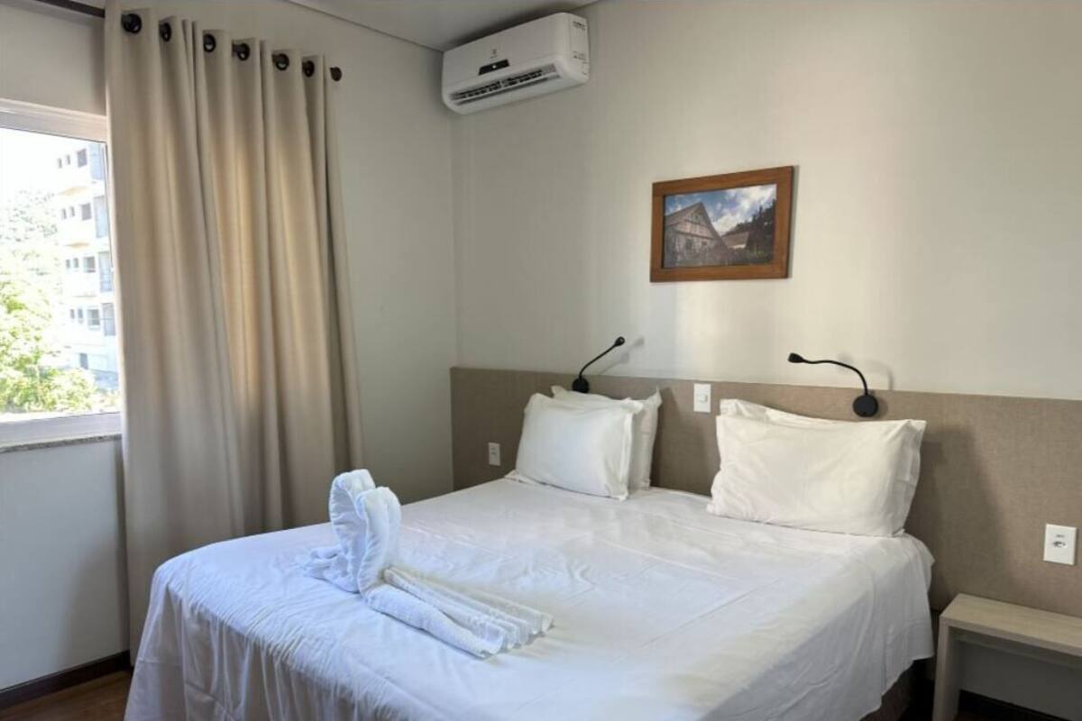 Habitación de hotel con una cama bien hecha, una mesita de noche, aire acondicionado instalado en la pared y un cuadro enmarcado.