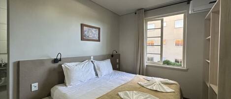 Ein Hotelzimmer mit Bett, Nachttisch, Fenster mit Vorhängen und einer Wand-Klimaanlage.