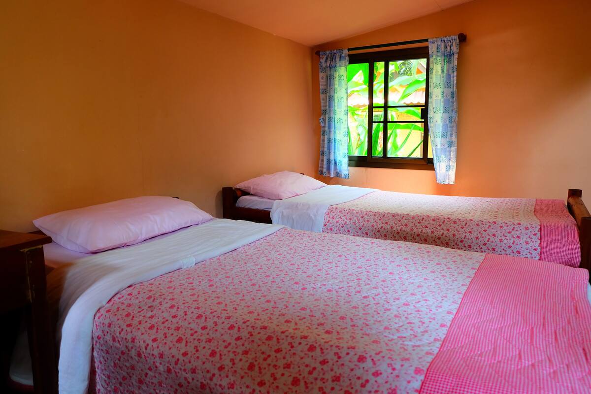 A room with two beds, each with a pink floral bedspread and a white pillow. There is a window with a green view outside.