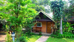 A small brick house with a sloped roof, surrounded by lush greenery and a concrete pathway leading to the entrance.