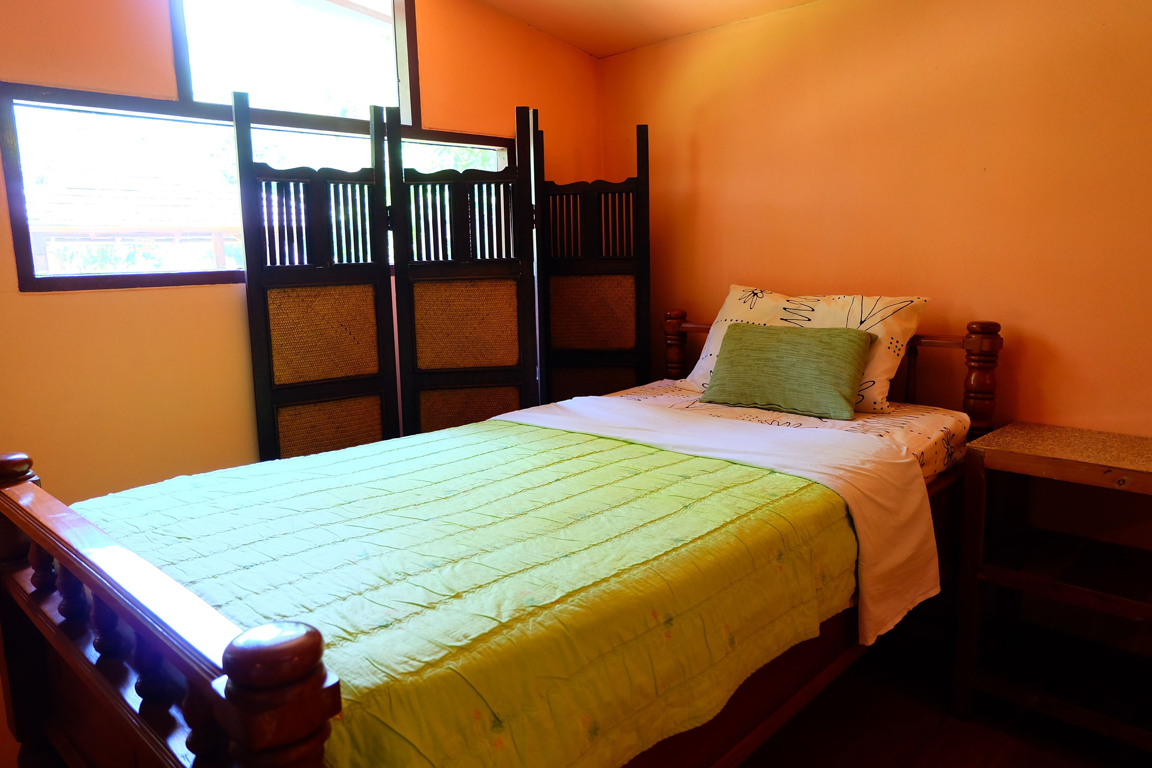 A single bed with a green quilt, a wooden headboard, a nightstand, and a window with blinds.
