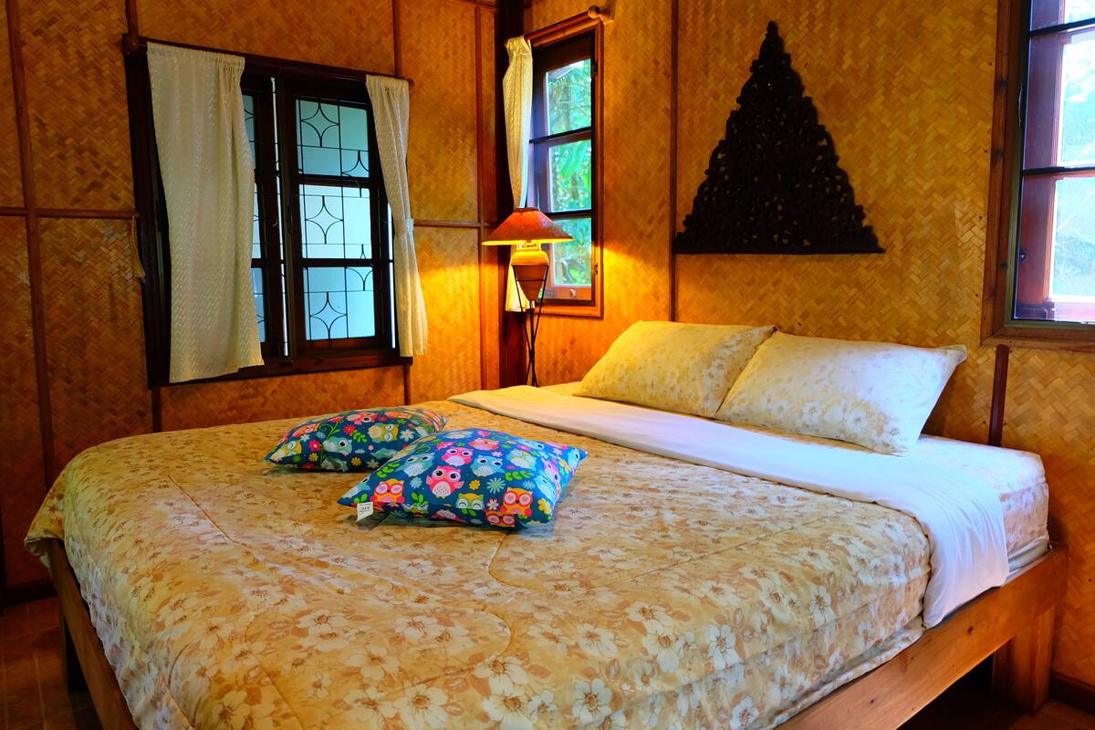 A bed with floral bedding, two pillows, a nightstand with a lamp, and a window with curtains.