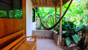 A wooden bench under a wooden pergola with a hammock, surrounded by lush green plants.