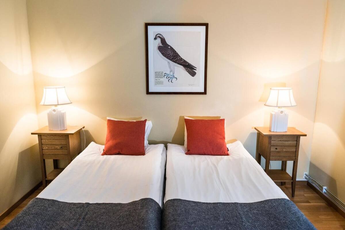 A double bed with white and grey bedding, two red pillows, and a framed bird illustration on the wall.