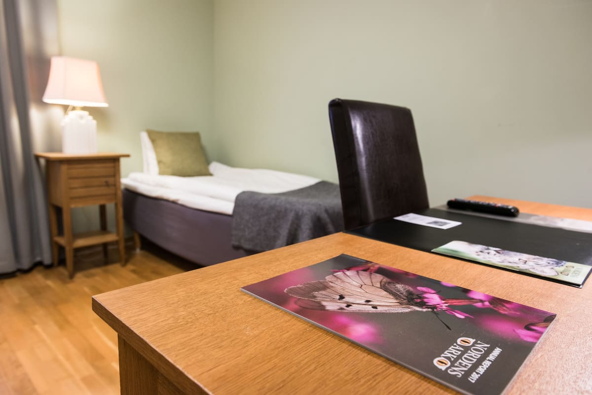 A hotel room with a bed, a wooden nightstand with a lamp, a desk with a laptop and a remote control, and a chair.