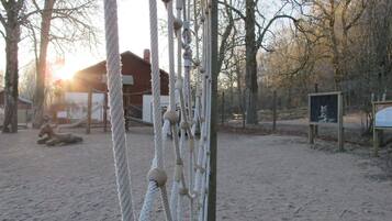 A rope climbing structure in a sandy area with trees and a wooden building in the background.