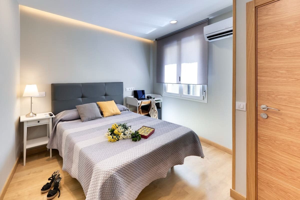 A modern bedroom with a bed, bedside tables, a desk, and an air conditioning unit.