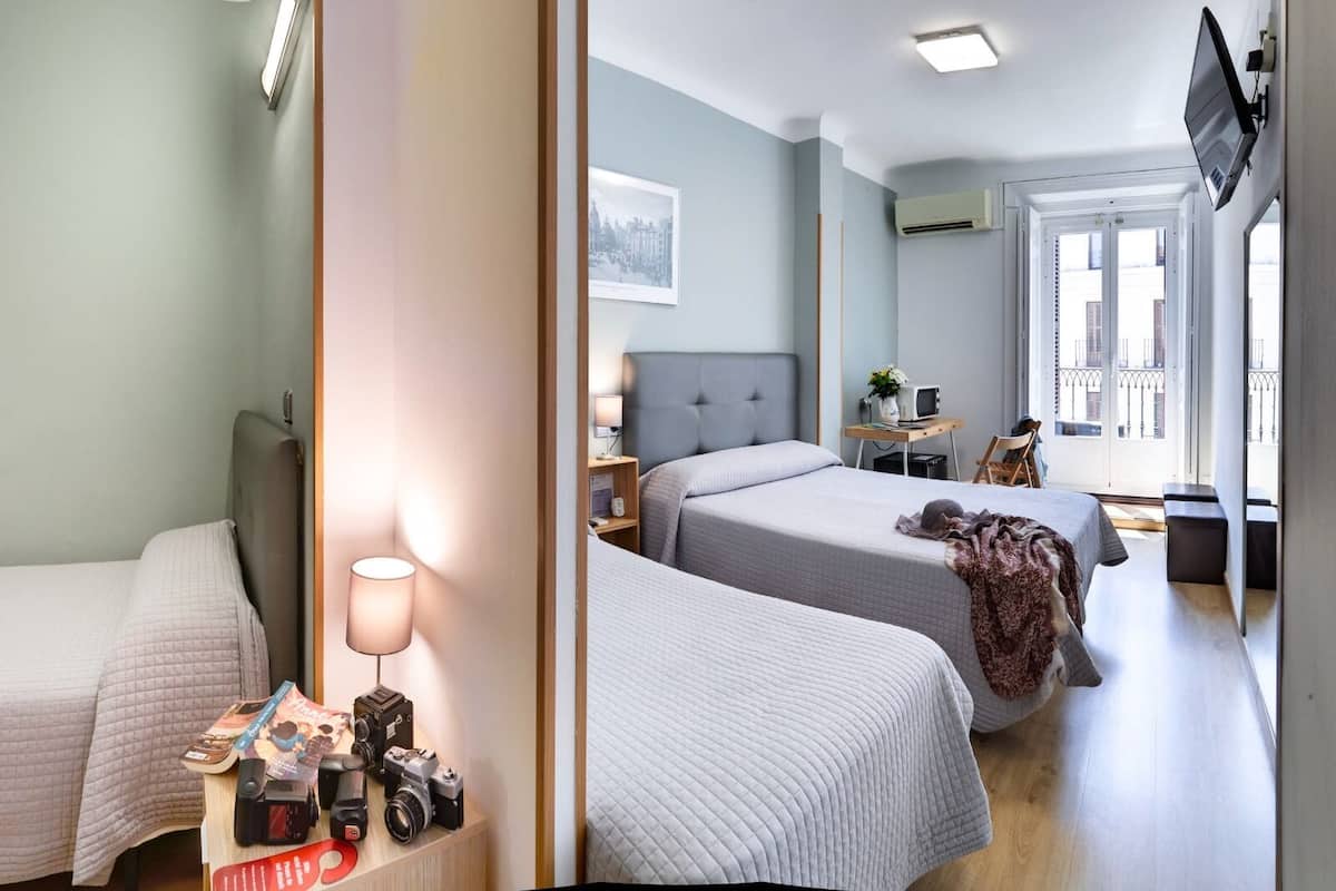 A hotel room with two beds, a desk with a lamp, a TV mounted on the wall, and a window with a view of buildings.