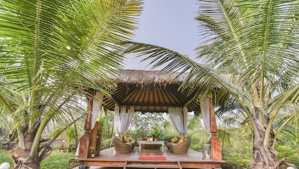 Un kiosque en bois avec un toit de chaume, entouré de palmiers, et comprenant un banc et une table avec des coussins.