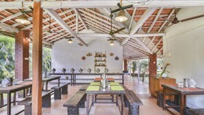 Un espace de restauration en plein air avec des tables et des bancs en bois, un mur de briques agrémenté d’objets décoratifs et un plafond avec des poutres apparentes et des luminaires suspendus.