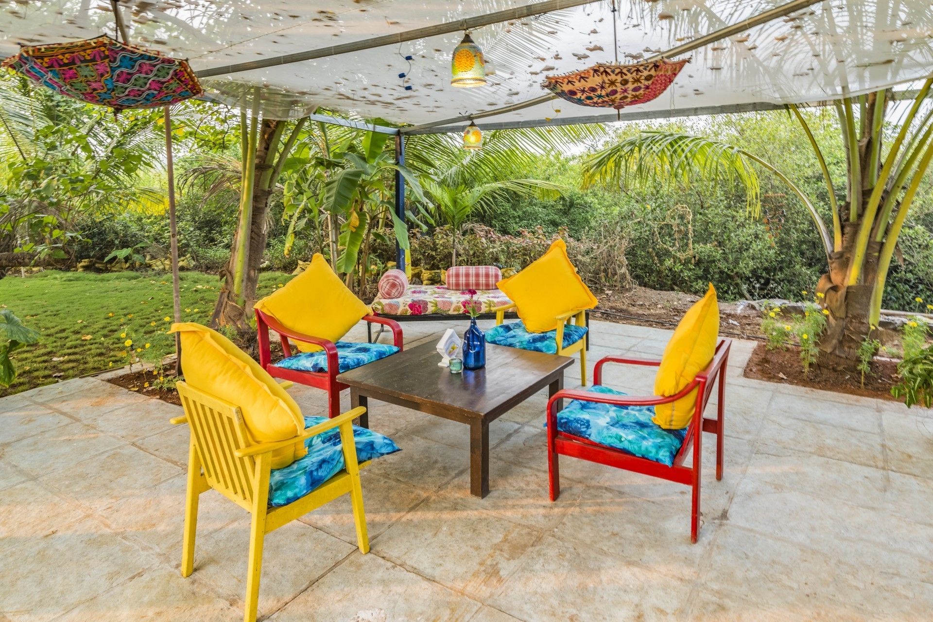Une terrasse extérieure avec une table et quatre chaises, entourée d’une végétation luxuriante et surmontée d’un auvent blanc.