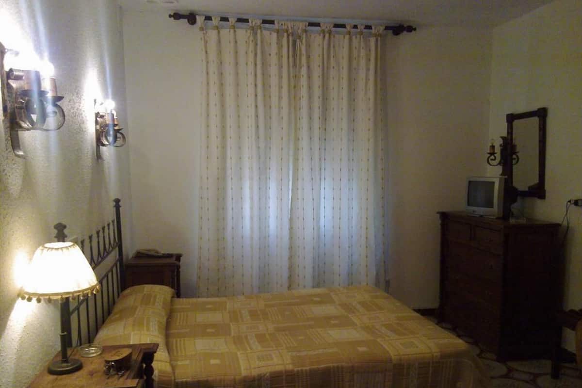 A bedroom with a bed, a nightstand with a lamp, a dresser, a television, and a window with curtains.