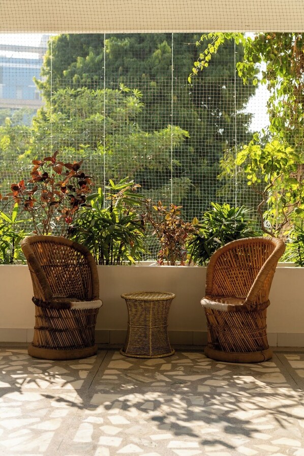 A sunlit outdoor seating area with wicker chairs and a small table, surrounded by lush greenery and a wire mesh barrier. - Girisadan - A City Centre Boutique Hotel (Jaipur)