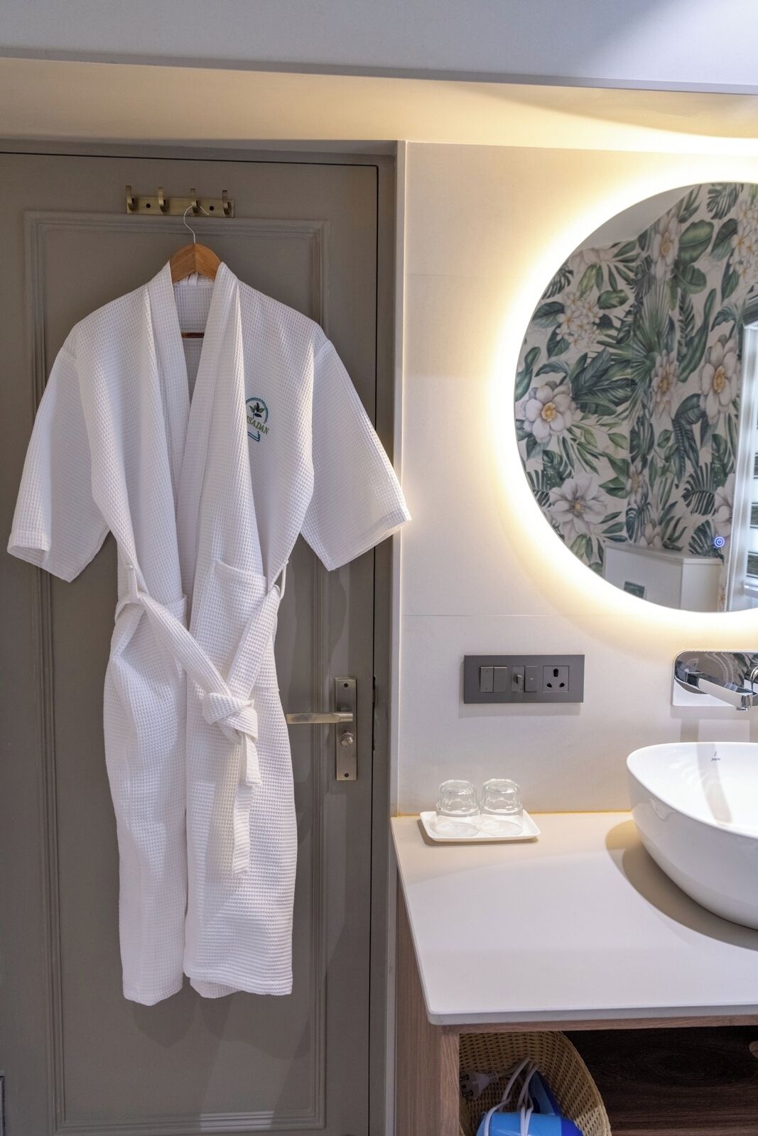 A bathroom with a white sink, a round mirror with a floral wallpaper, and a white robe hanging on a door.