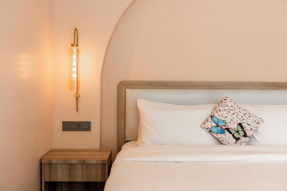 A neatly made bed with a white pillow and a decorative pillow with a butterfly pattern. There is a bedside table with a wooden finish and a wall-mounted light fixture.