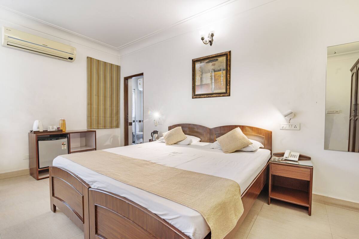 A hotel room with a wooden bed, a bedside table with a phone, a small desk with a coffee maker, and a wall-mounted air conditioner.