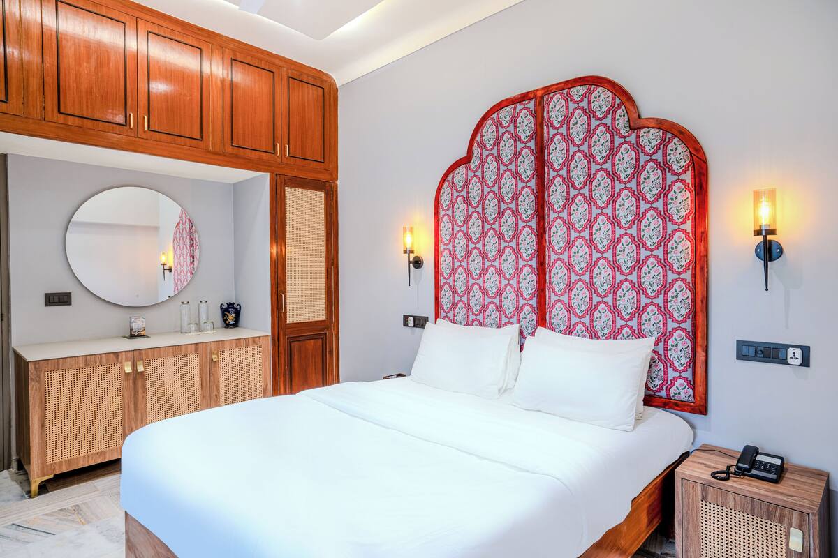 A neatly arranged bedroom with a large bed, wooden wardrobe, a round mirror, and patterned headboard.