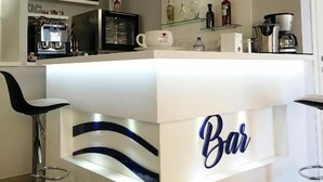 A bar area with a white counter, shelves with bottles, and two bar stools.