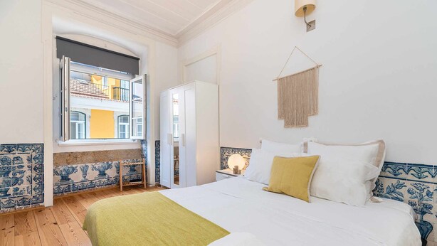 Una habitación de hotel con una cama, un ventanal con vistas a un edificio, un armario y una mesita de noche con lámpara.
