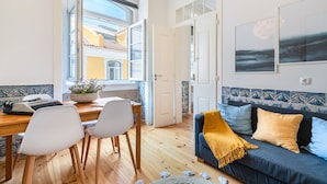 Una habitación con piso de madera, una mesa de comedor con sillas, un sofá con cojines, una planta en maceta y una ventana con puerta abierta.