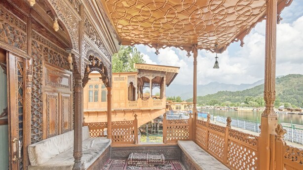 An ornate wooden balcony with intricate carvings, a patterned rug, and a view of a river and mountains. - GuestHouser 3 BHK Houseboat d520 (Srinagar)
