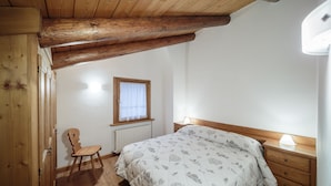 A bedroom with a wooden ceiling, a single bed with a patterned duvet, a bedside table with a lamp, a chair, and a window with a white curtain. - Villa Lacedel (Cortina d'Ampezzo)