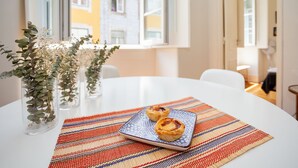 A table with two pastries on a patterned placemat, a vase with greenery, and a view of buildings through a window. - Gonzalo's Guest Apartments - Downtown Historic Flats I (Lisbon)