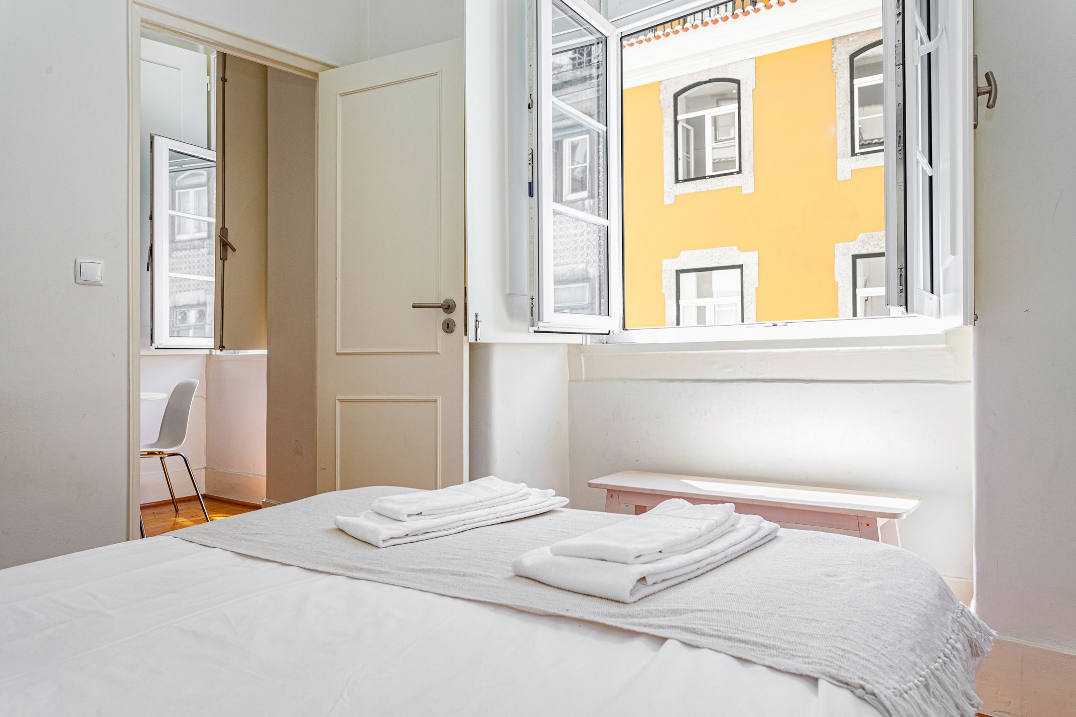 A hotel room with a bed, a chair, a window, and a view of a yellow building.