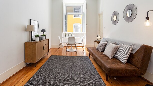 A modern living room with a brown sofa, a wooden cabinet, a dining table with chairs, and a large window with a view of a yellow building. - Gonzalo's Guest Apartments - Downtown Historic Flats I (Lisbon)