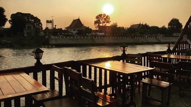 Un restaurante a orillas del río con mesas y sillas de madera, vistas al atardecer sobre el agua y edificios tradicionales al fondo.