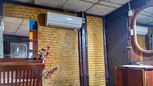 Una habitación con un banco de madera, una mesa pequeña con un florero y aire acondicionado de pared, y una puerta de madera con un espejo.