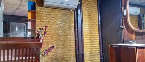 Une pièce avec un banc en bois, une petite table sur laquelle est posé un vase de fleurs, un climatiseur fixé au mur et une porte en bois dotée d’un miroir.