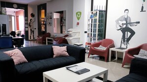 A modern living room with a black sofa, a white coffee table, and red chairs. There is a mural of a man playing a guitar on the wall. - Alternative Creative Youth Hostel Barcelona (Barcelona)