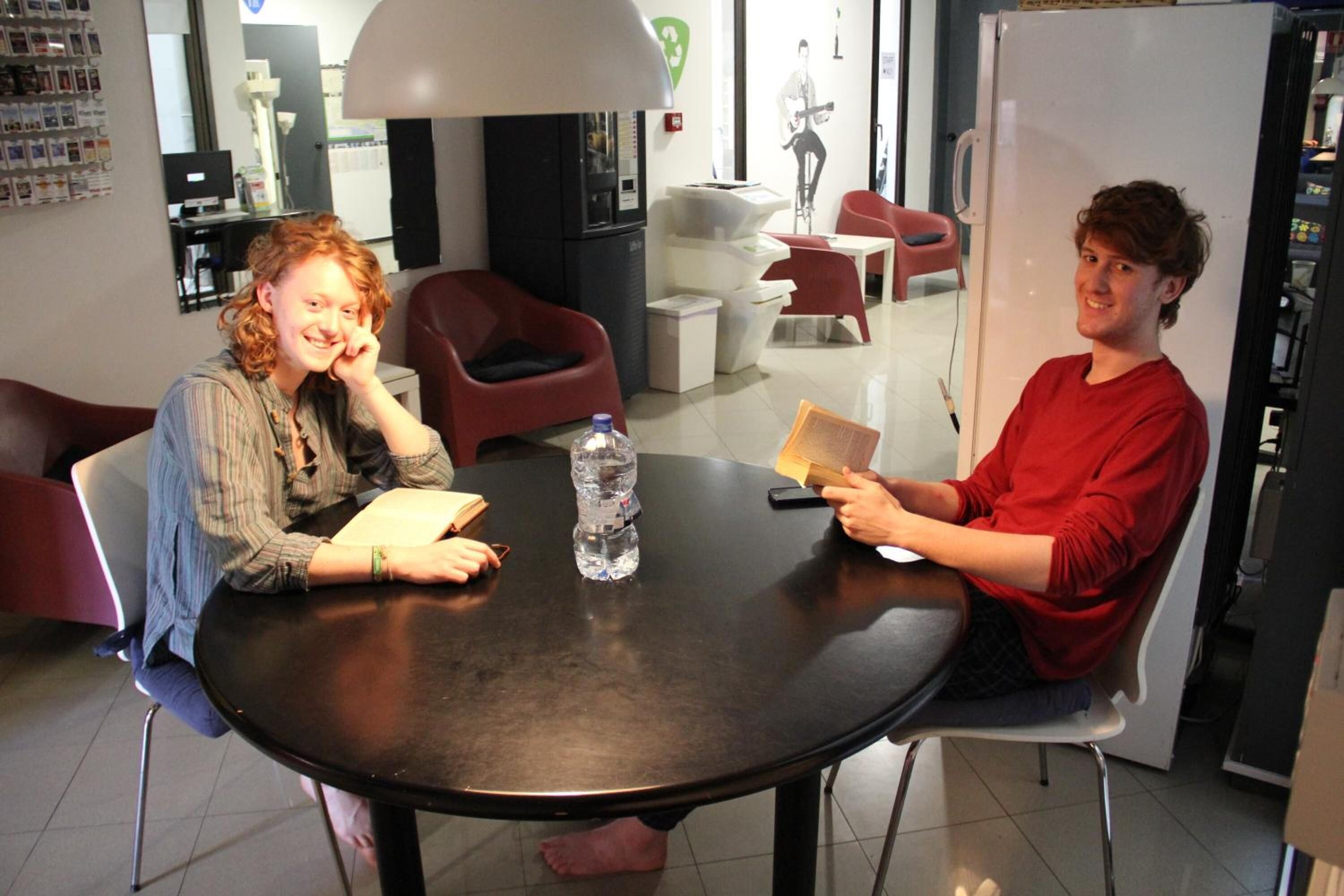 Two people seated at a round table with a water bottle and papers in a modern room with chairs and a refrigerator.