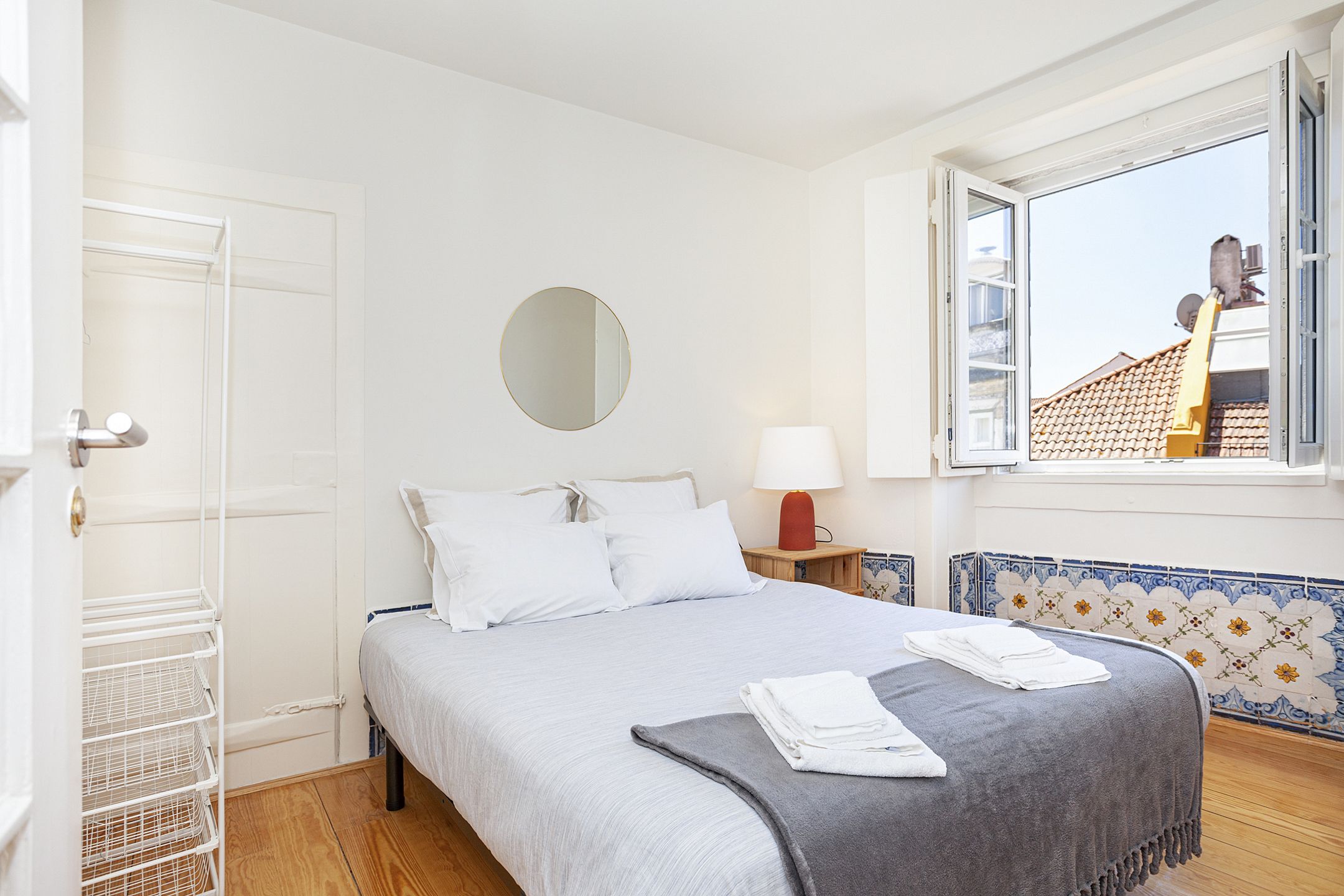 A bedroom with a bed, a round mirror, a nightstand with a lamp, a window with a view of buildings, and a tiled wall.